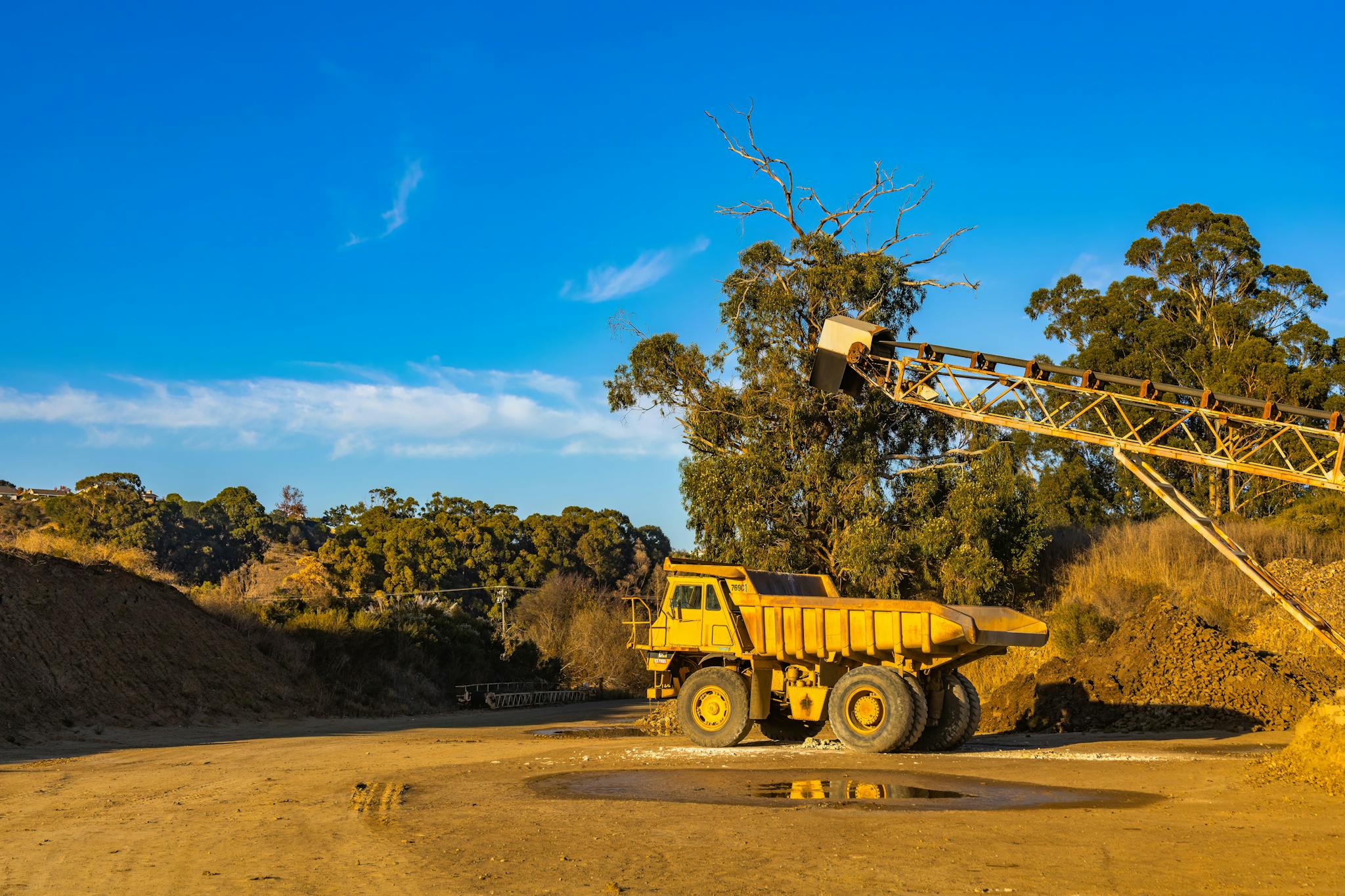 A large dump truck and conveyor in a quarry under a bright blue sky.